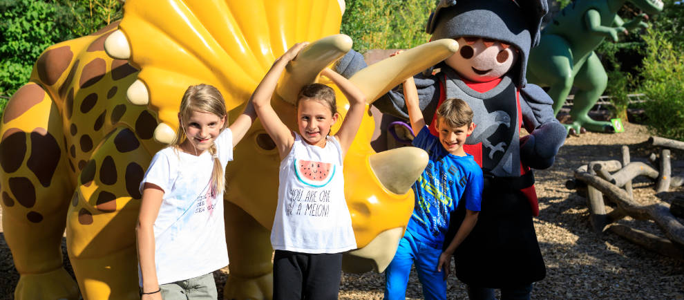 Eine Gruppe von lachenden Kindern steht vor einem gelben Dinosaurier. Mit dabei ist eine lebensgroße Playmobilfigur im Ritterkostum.